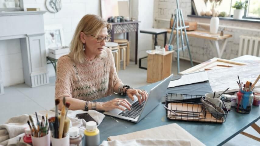 Your Own eCommerce Store An elderly artist in her studio, using a laptop to learn how to sell art online.