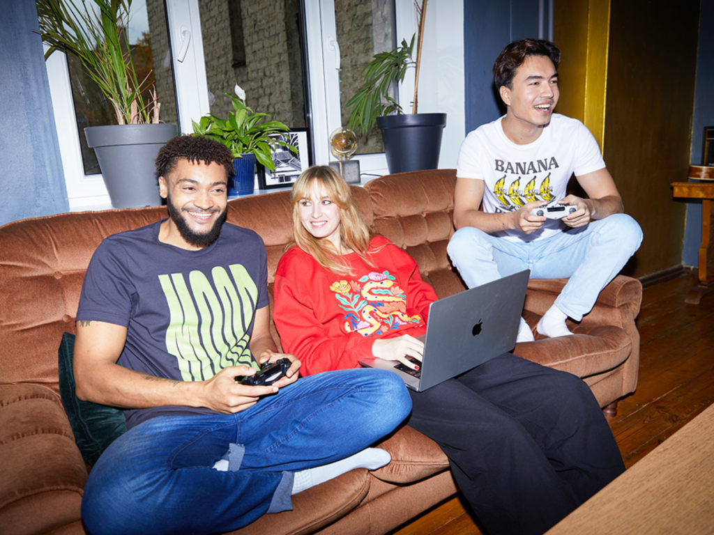 Start your weekend business with Printify A group of people sitting on a couch, playing video games, wearing custom t-shirts and a hoodie.